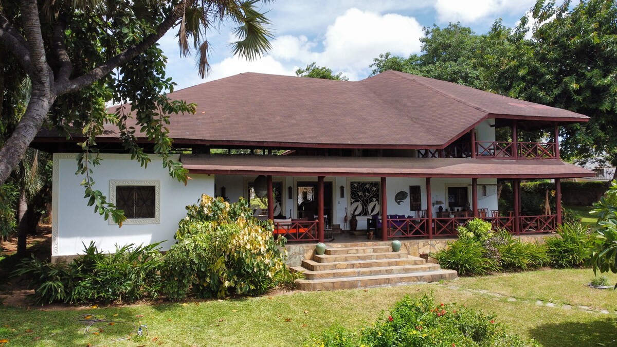 A two-story villa is surrounded by lush greenery, featuring a broad outdoor patio with several steps leading to the entrance. Shaded by overhanging trees, the structure includes a spacious balcony with red railings, providing views of the tranquil garden and inviting spaces beyond.