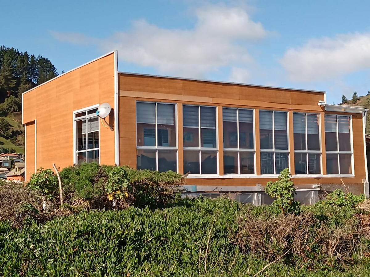 A modern house is presented with a wooden exterior, featuring large windows that allow for ample natural light. Lush greenery surrounds the base, and a clear blue sky can be seen above, creating a serene backdrop.