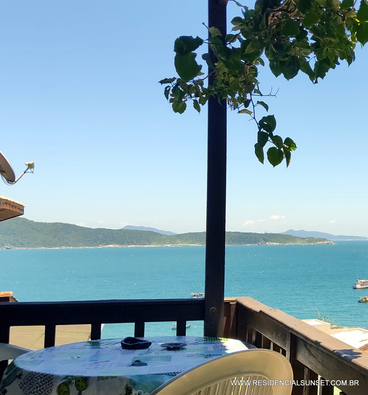 A private balcony view features a light-colored table set with amenities, overlooking a vibrant blue sea. In the distance, green hills are visible, complementing the tranquil waters. A clear sky enhances the scenic panorama, creating a peaceful outdoor setting.