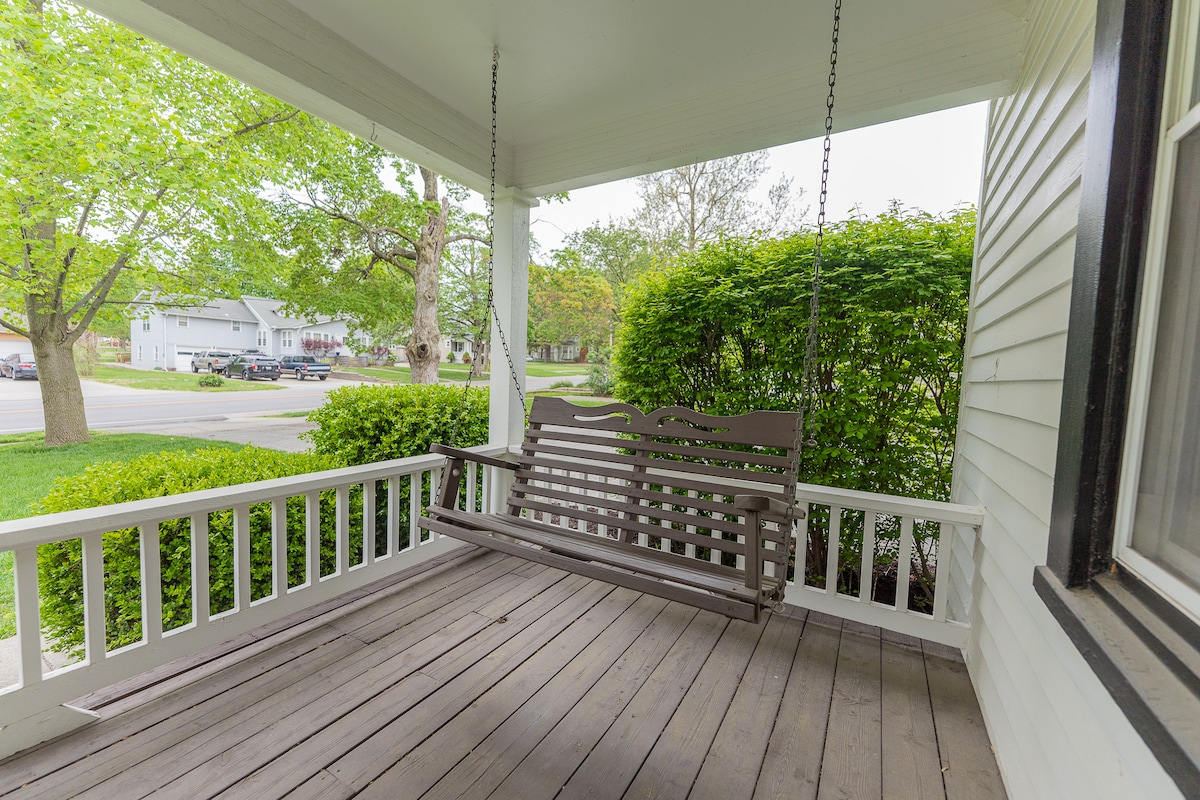 A wooden swing is positioned on a covered porch, inviting relaxation. Lush greenery frames the view, with a glimpse of the street beyond. The porch features wooden flooring and a railing painted white, contributing to an open and welcoming outdoor space.