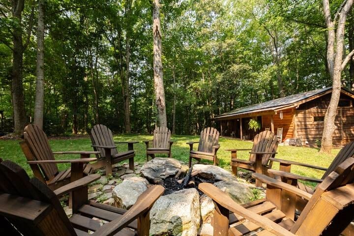 A fire pit surrounded by six wooden Adirondack chairs sits on a grassy area, framed by tall trees. The nearby cabin is partially visible, with a sloped roof and a rustic wooden facade, offering a cozy outdoor gathering space.