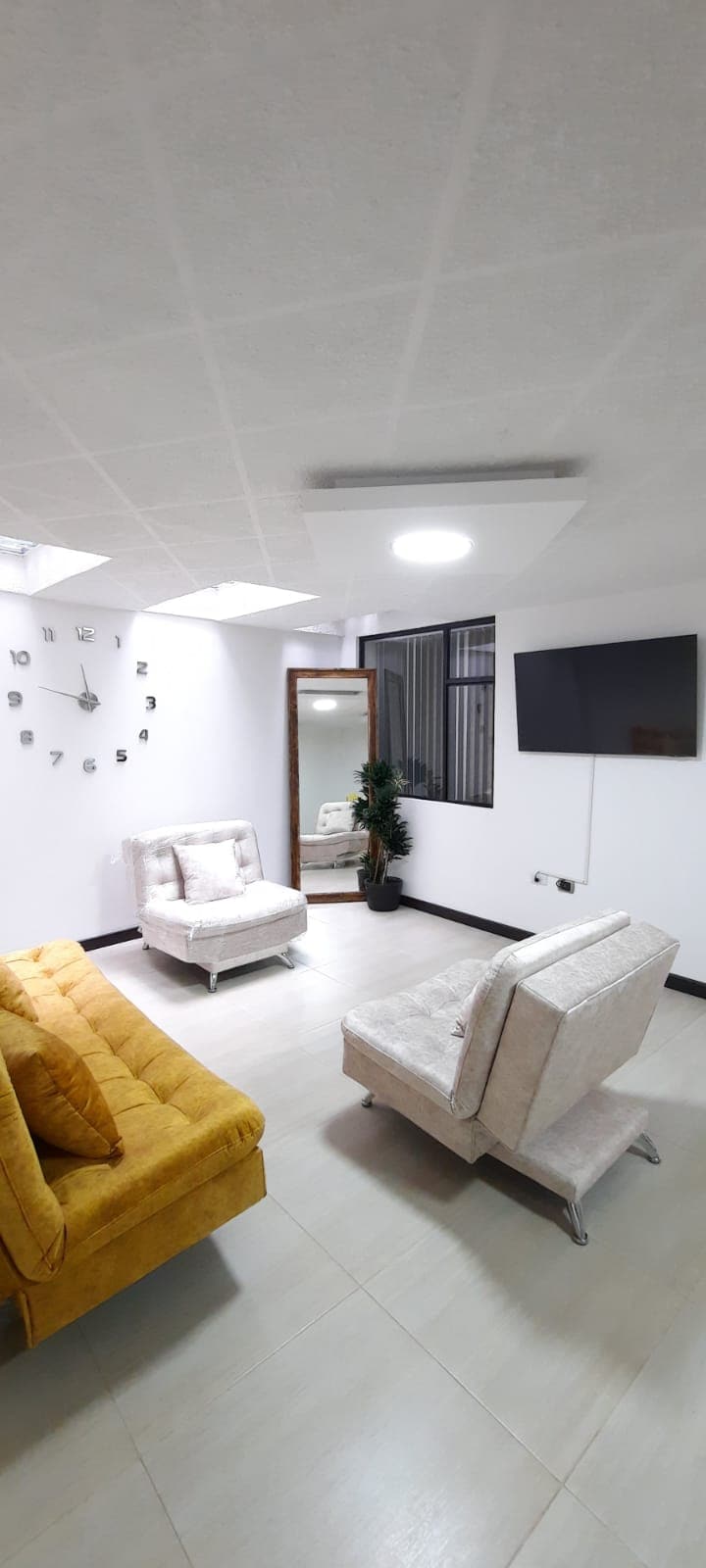 A well-lit living area features a yellow sofa and two light-colored armchairs, creating a comfortable seating arrangement. A large mirror and a decorative plant enhance the space, while a wall clock adds a functional touch. A mounted television is also present on the wall.