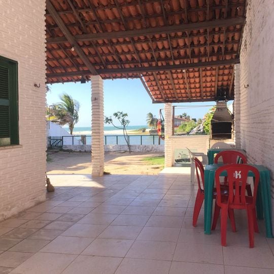 A covered patio area features a table with four colorful chairs, offering a view of the ocean in the distance. The space is framed by white brick columns and a ceiling with rustic tiles, providing a relaxed outdoor atmosphere.