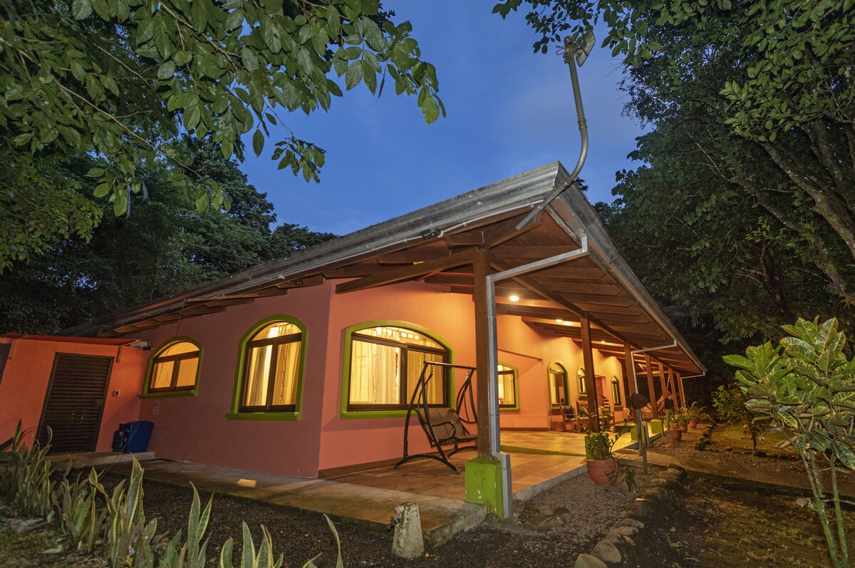 The exterior of Bird Paradise is shown at twilight, with warm lights glowing through the arched windows. The vibrant pink facade is complemented by green trim. A covered porch features outdoor seating, surrounded by lush vegetation and trees creating a peaceful ambiance.