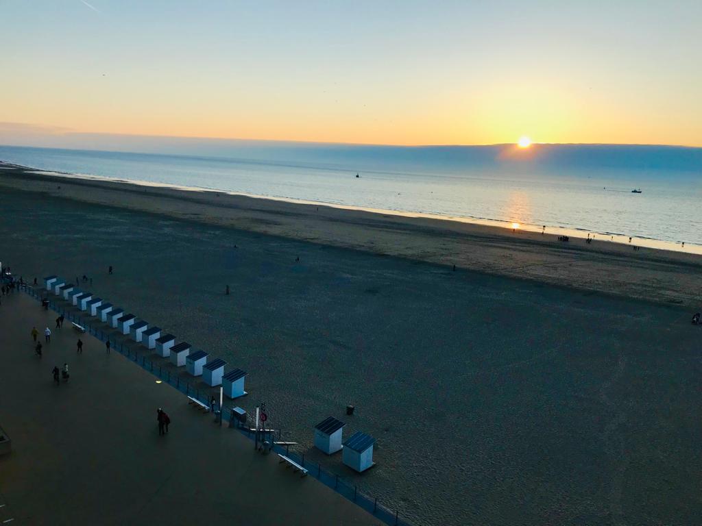 A serene beach view is captured at sunset, with a calm sea reflecting soft hues of orange and blue. A stretch of sandy shoreline is visible, lined with beach cabins, while small figures are seen walking along the water's edge.
