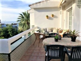 An outdoor terrace is featured, furnished with a dining table and chairs. Decorative plants are placed along the white wall. Light cushions are arranged on a lounge area, inviting relaxation while overlooking greenery and a glimpse of the sea in the background.