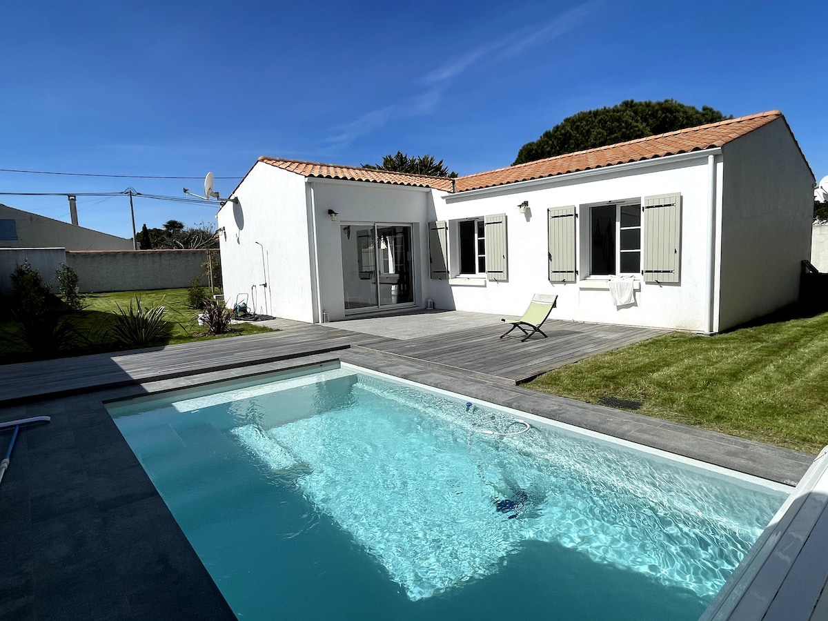 A charming house is featured alongside a heated swimming pool. The surrounding wooden deck offers a sun lounger, while the grassy area adds greenery. The light-colored exterior is complemented by green shutters, and large glass doors lead into the inviting interior.