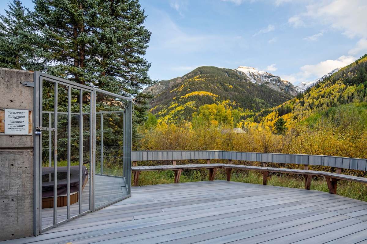 A spacious outdoor deck features a circular hot tub surrounded by a curving bench, all set against a backdrop of colorful autumn foliage and mountains. The area is enclosed by a glass gate, allowing unobstructed views of the natural landscape.