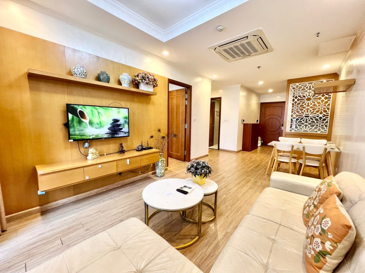A light-filled living area features a sleek wooden entertainment unit with a flat-screen TV. A comfortable sofa is arranged around a round coffee table, adorned with decorative items. In the background, a dining table for four is positioned next to a bright window.