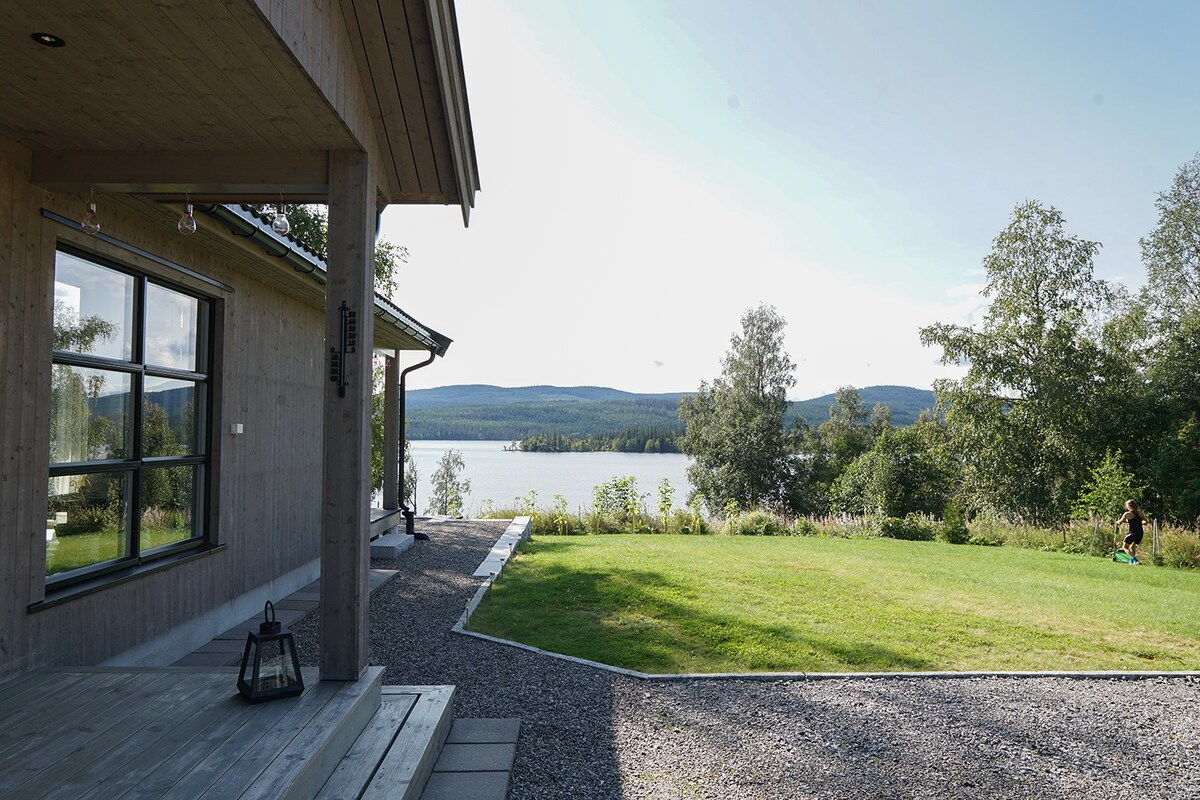 A modern wooden cabin is positioned near a serene lake, with expansive views of the surrounding landscape. A well-maintained lawn extends from the cabin towards the water, while gravel paths and outdoor seating areas enhance the inviting exterior space.