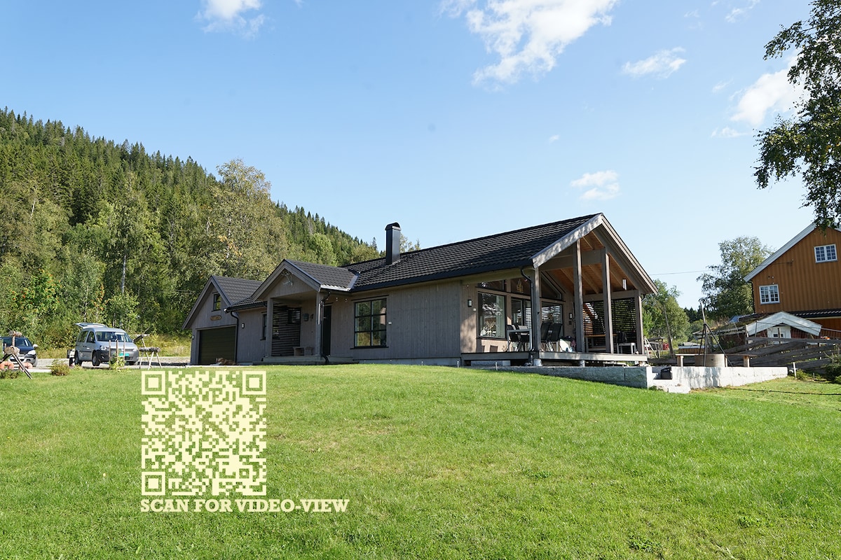 A spacious cabin is situated on a green lawn, framed by trees and hills. Large windows and a covered patio are visible, creating a welcoming outdoor space. A gravel path leads to the entrance, and neighboring buildings are faintly seen in the background.