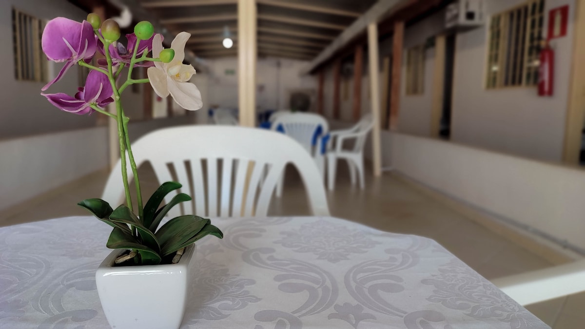 A small white vase holds a delicate arrangement of purple and white orchids, placed on a table adorned with a light-colored tablecloth. The background reveals a spacious area with white chairs and soft lighting, creating a calm and inviting atmosphere.