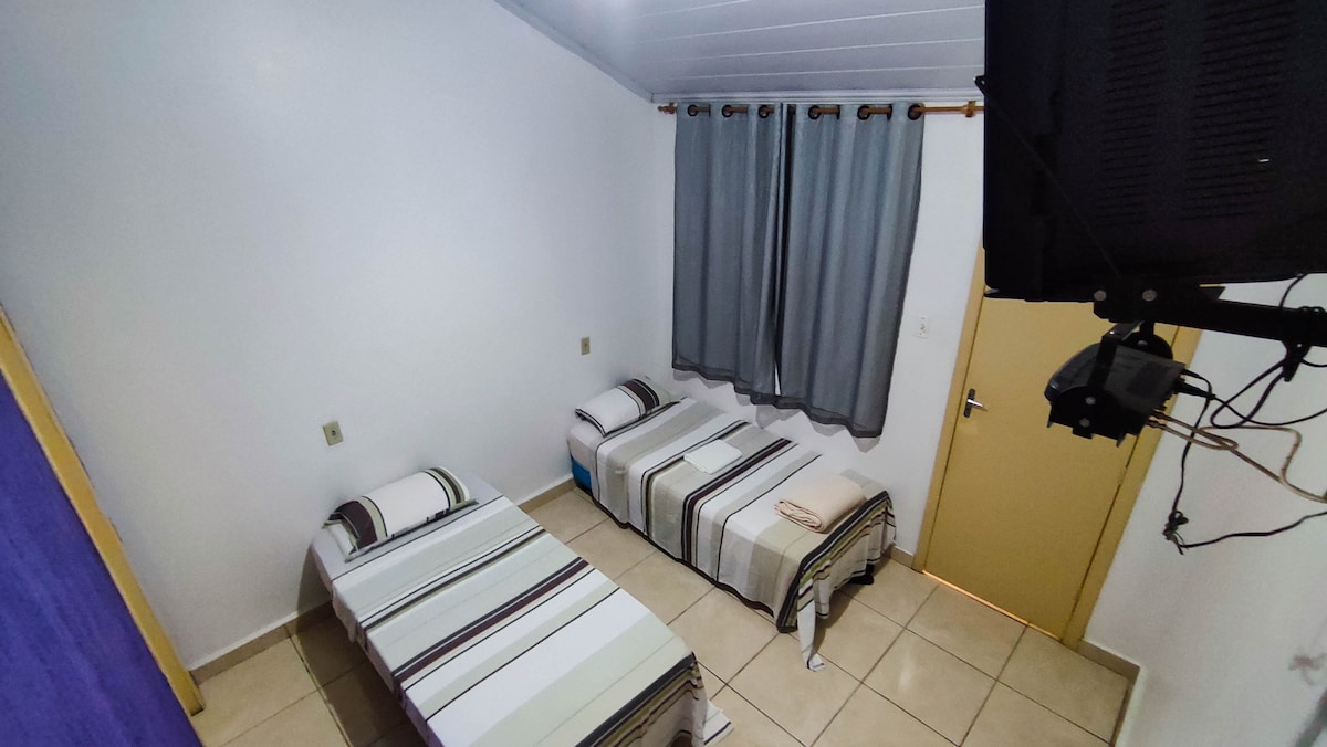 The room features two twin beds with striped bedding, positioned against a light-colored wall. A gray curtain covers the window, and a wall-mounted TV is visible in the corner. The tiled floor adds to the simple, clean aesthetic of the space.