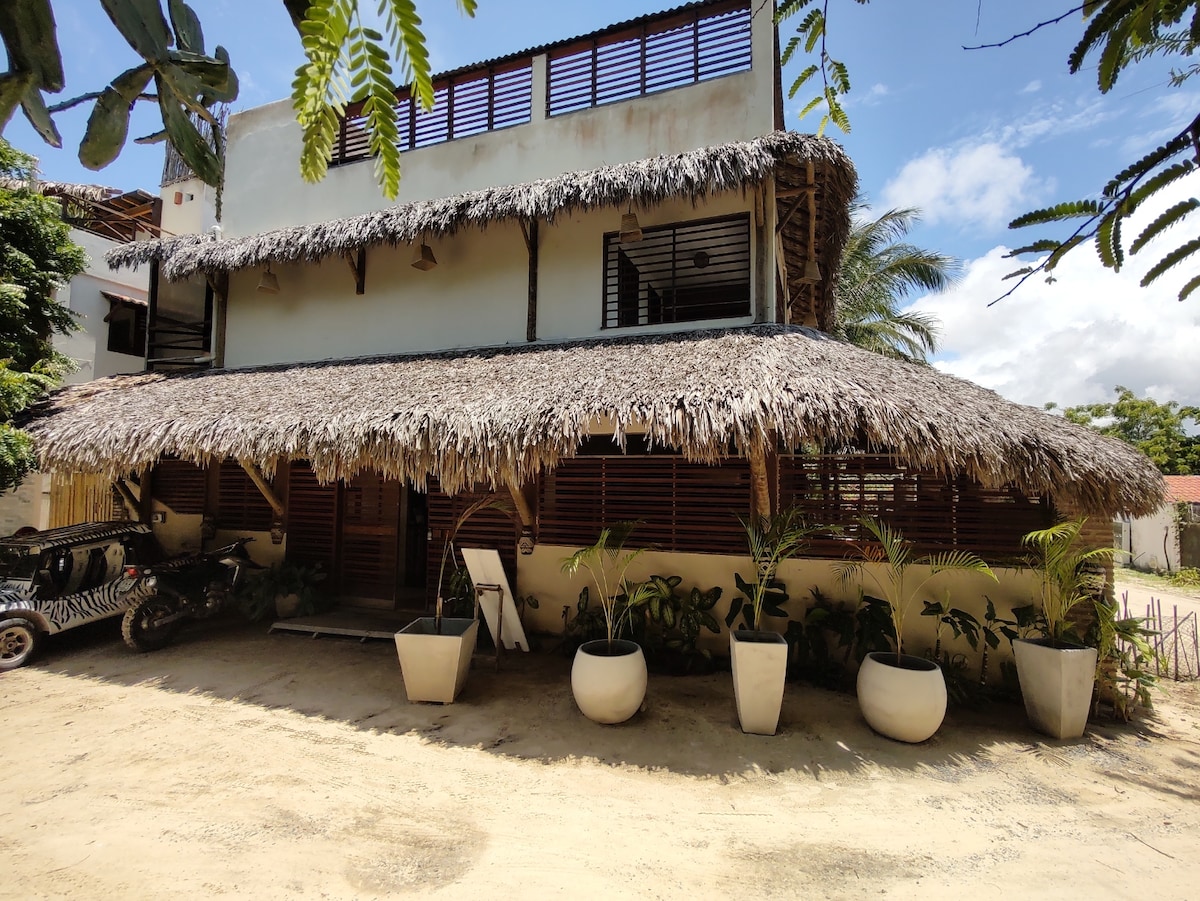 A rustic building is presented with a thatched roof, blending naturally with its surroundings. Large planters house lush greenery at the entrance. The structure features wooden slatted windows and a spacious ground level, contributing to a welcoming outdoor atmosphere.