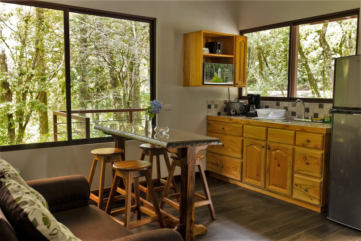 A bright dining area is enhanced by large windows offering forest views. The kitchen features wooden cabinetry and modern appliances, including a refrigerator and coffee maker. A wooden table with three stools complements the cozy seating area, creating an inviting space for meals and relaxation.