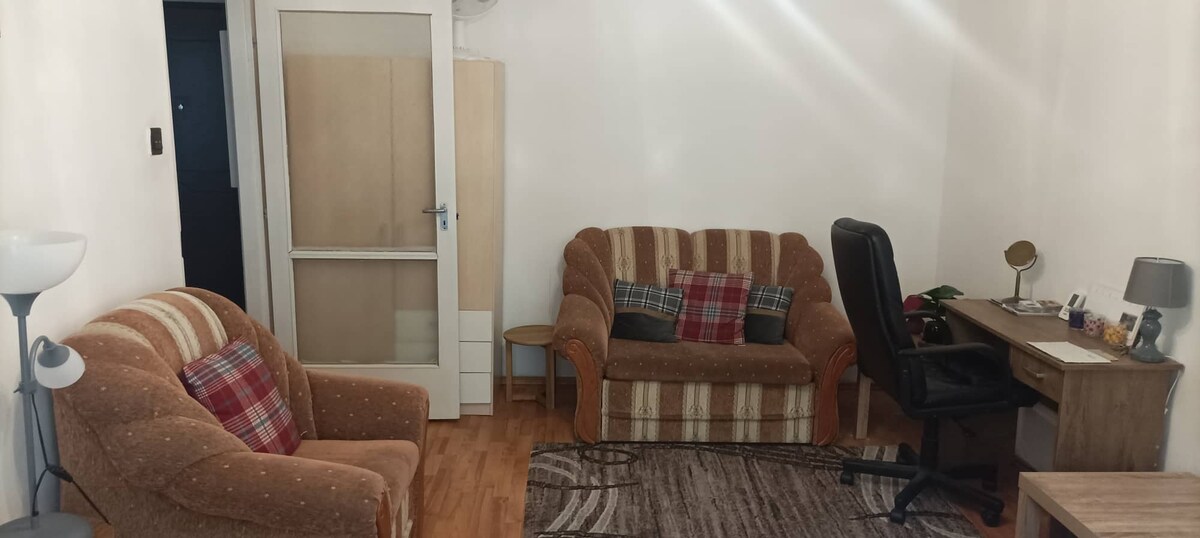 A cozy living room area includes two armchairs and a sofa adorned with throw pillows. A small table and desk with a chair are positioned next to a wardrobe. The wooden flooring and a patterned area rug contribute to the comfortable setting.