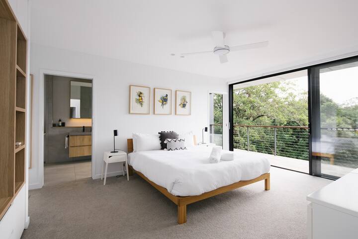 Master bedroom with King sized bed, Smart TV, block-out blinds, plush carpet and private balcony looking out to nature. The ensuite can be seen on the left.