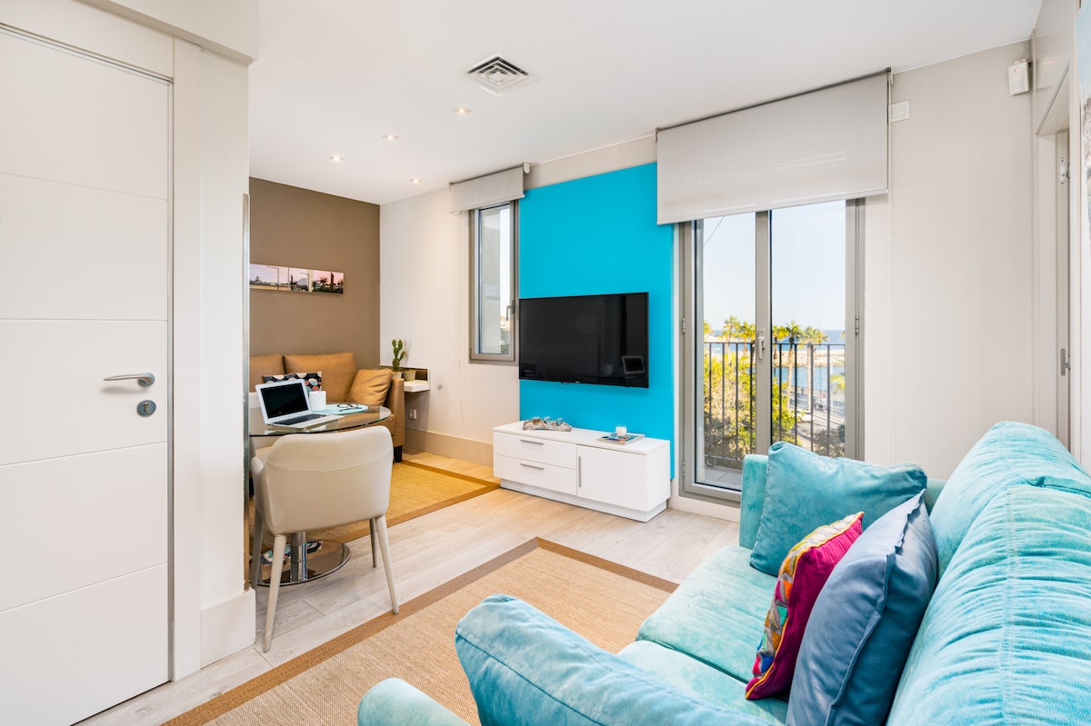 A modern living area features a vibrant blue accent wall and large windows allowing natural light to pour in. Comfortable seating is arranged around a sleek white television unit, with a small dining space visible in the background and an inviting ambiance created by warm, neutral tones.