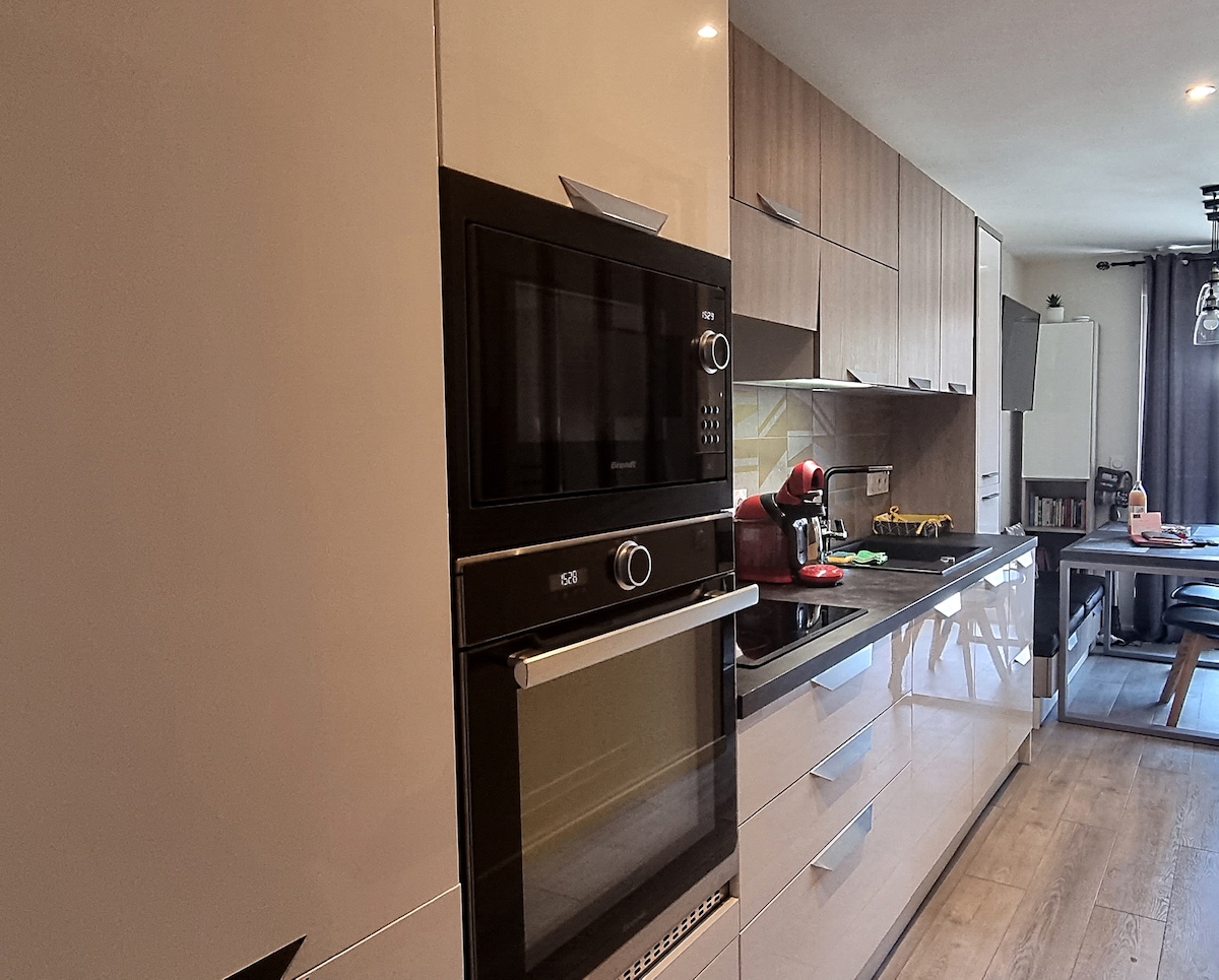 A modern kitchen features sleek cabinetry with a mix of light and dark finishes. Essential appliances include a built-in oven and microwave, a coffee machine, and a stovetop. A dining table with chairs is positioned nearby, providing a functional and stylish space for meal preparation and dining.
