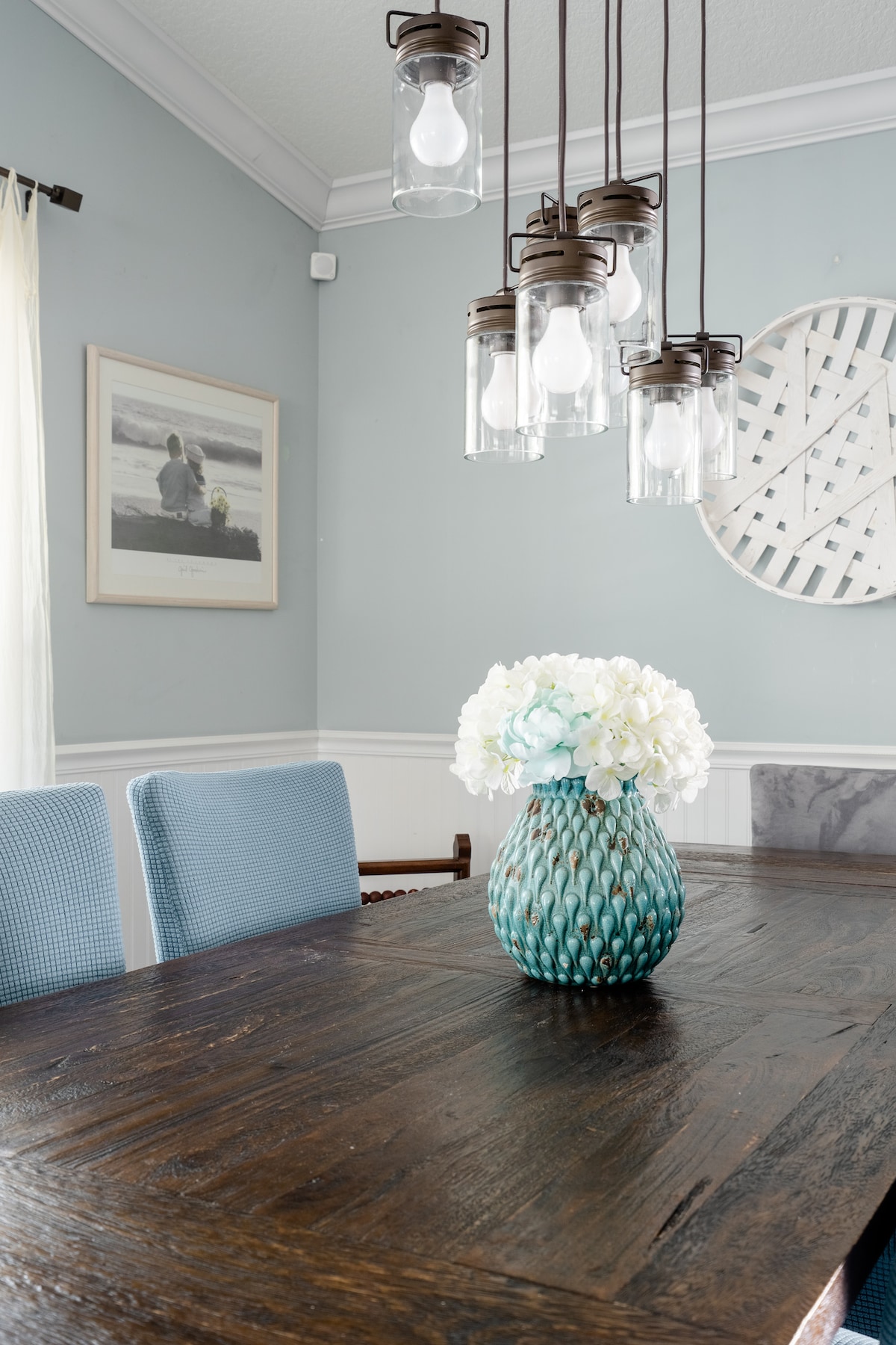 A rustic wooden dining table is positioned beneath an elegant light fixture with hanging glass bulbs. A teal decorative vase filled with white flowers serves as the centerpiece. Soft blue chairs complement the light blue walls, enhancing the room's tranquil ambiance.