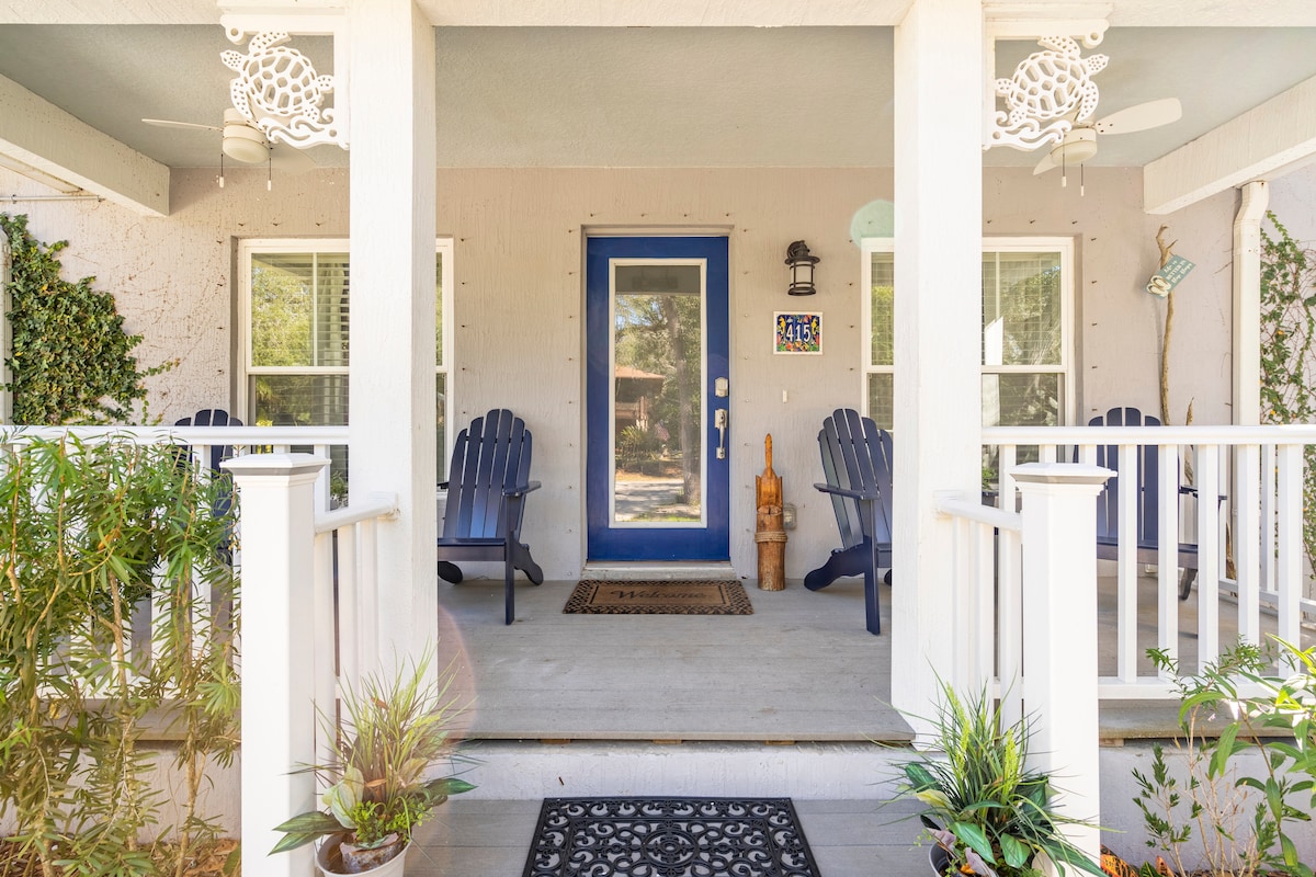 The quaint front porch features two rustic adirondack chairs, inviting relaxation. Potted plants are arranged on both sides of the entrance, while a doormat lies at the entry. A blue front door is accented by a small decorative piece, creating a welcoming entryway.