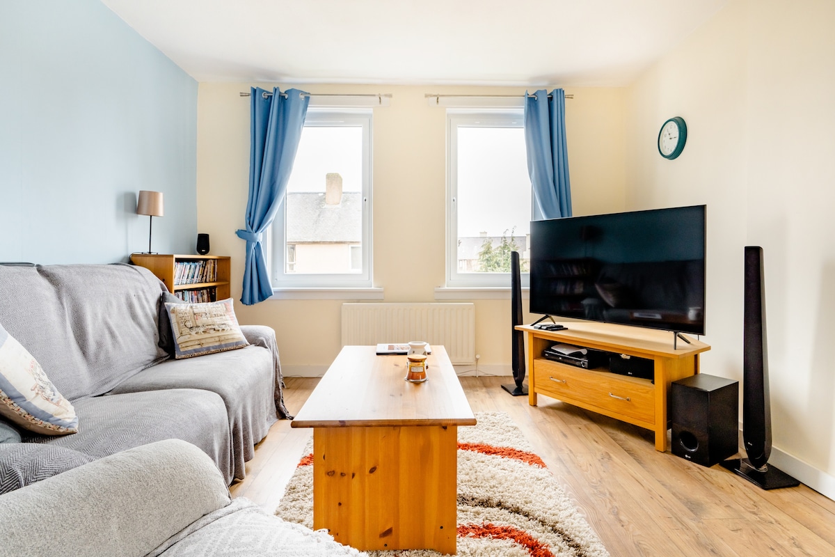 The living room features two windows with light blue curtains, allowing natural light to fill the space. A comfortable sofa sits alongside a wooden coffee table. A large TV is mounted on a stand, with speakers positioned on either side, creating an inviting area for relaxation.