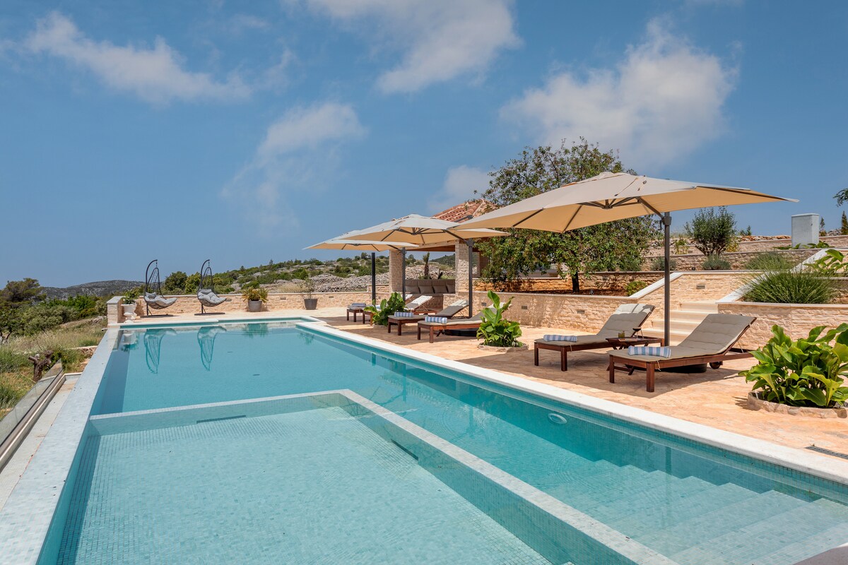 A large swimming pool reflects the clear sky, surrounded by elegant lounge chairs and shaded by large umbrellas. Lush greenery and low stone walls create a calming outdoor space that invites relaxation.