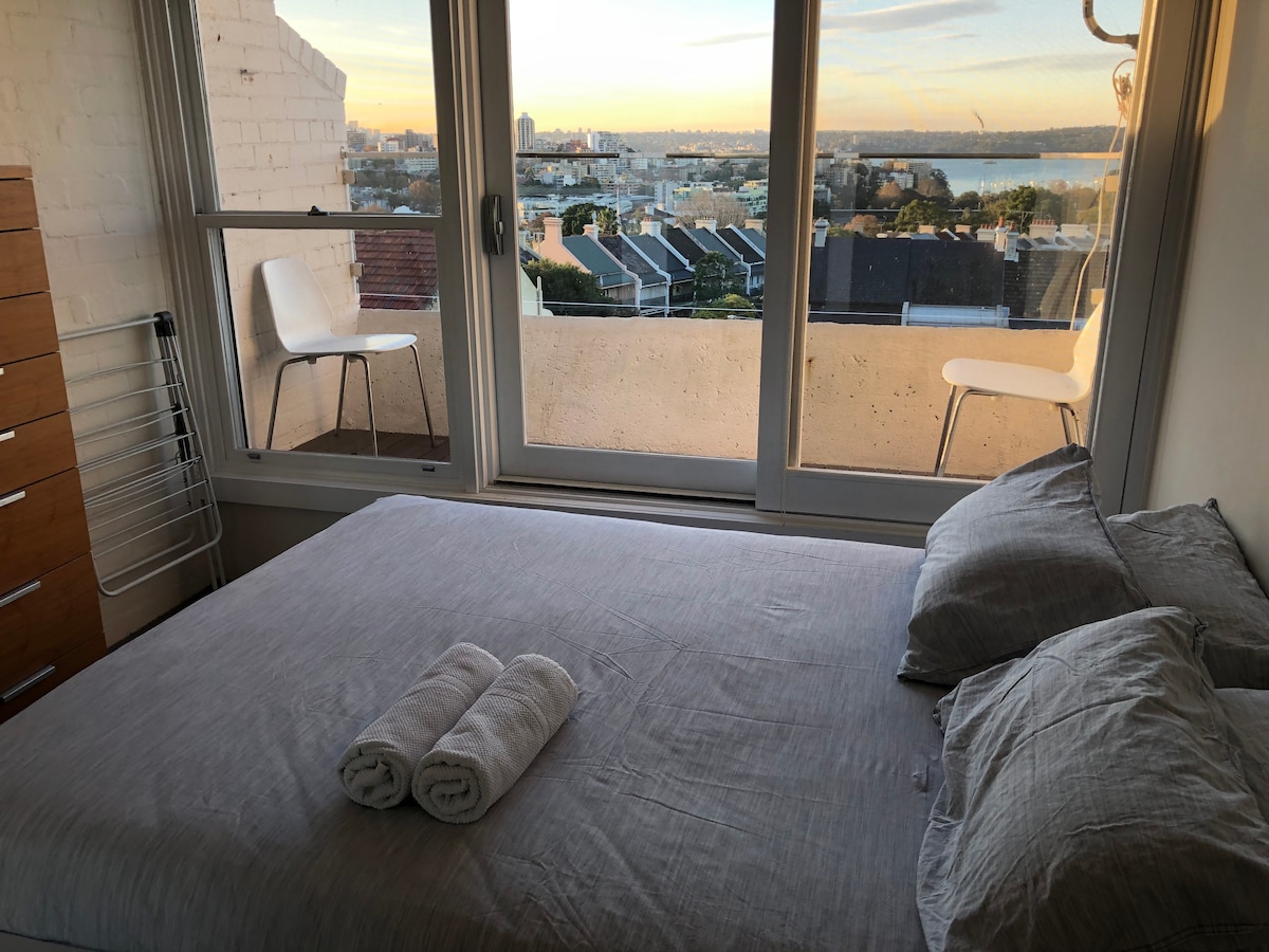 A cozy bedroom features a spacious bed with neatly arranged white towels at the foot. Large glass doors open to a balcony, revealing views of the city and harbour. A wooden dresser and two minimalist chairs enhance the tranquil space.