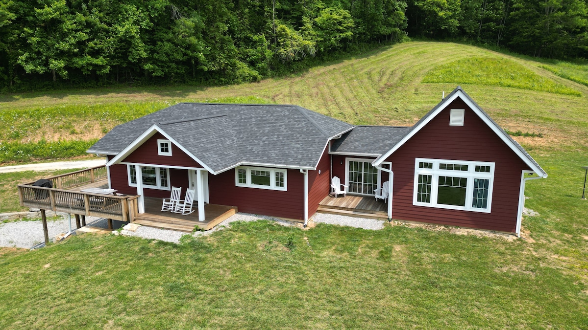 A charming home with a reddish exterior is surrounded by lush greenery and expansive fields. A wooden deck with rocking chairs is visible in the front, while large windows provide natural light to the interior. The property features a spacious lawn leading up to the house.