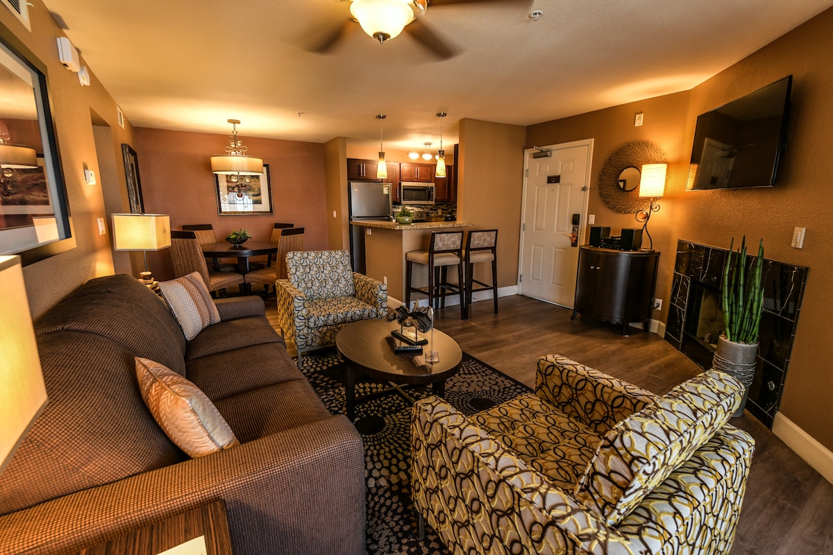 An open-concept living area features a comfortable sofa, two patterned armchairs, and a coffee table, all arranged around a cozy fireplace. A flat-screen TV is mounted on the wall. The adjoining kitchen area is visible, highlighting a bar with stools and a dining space.