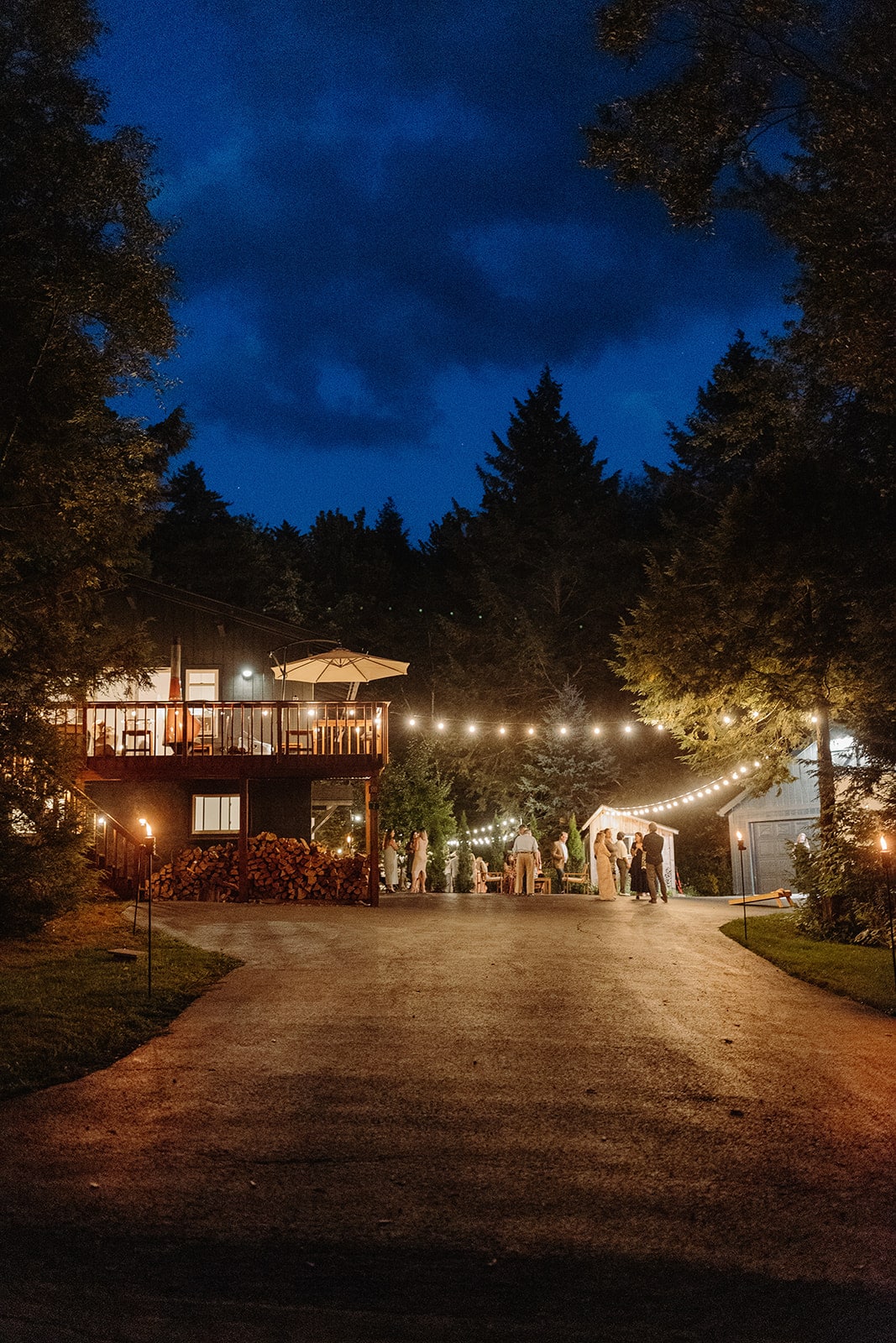 A modern chalet is illuminated at dusk, surrounded by trees. String lights create a warm ambiance over the outdoor gathering area. Guests can be seen enjoying the space on the driveway leading to the home, enhancing the inviting atmosphere of the location.