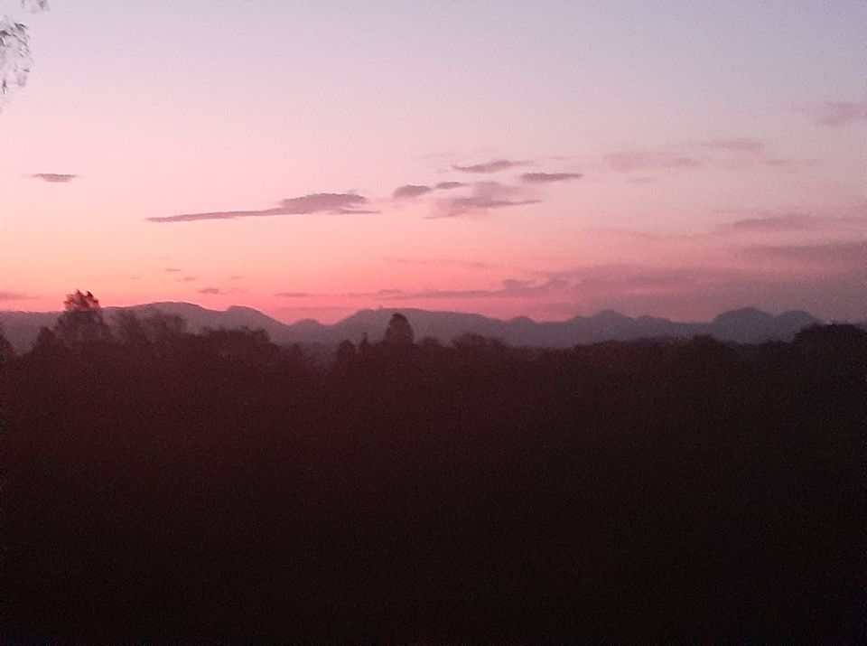 A serene twilight view showcases a gradient of soft pinks and purples in the sky, gently illuminating the distant Warrumbungle mountains. Shadows of surrounding bushland create a tranquil foreground, contributing to the peaceful atmosphere of the landscape.
