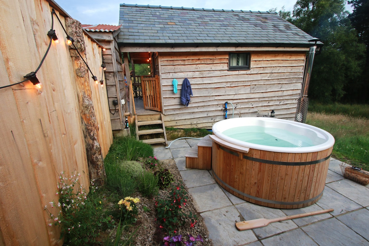 A secluded wood-fired hot tub is situated next to a rustic cabin, surrounded by a tasteful stone patio and vibrant flowers. Warm evening light enhances the natural wood siding of the cabin, while steps lead up to its entrance, inviting guests to relax.