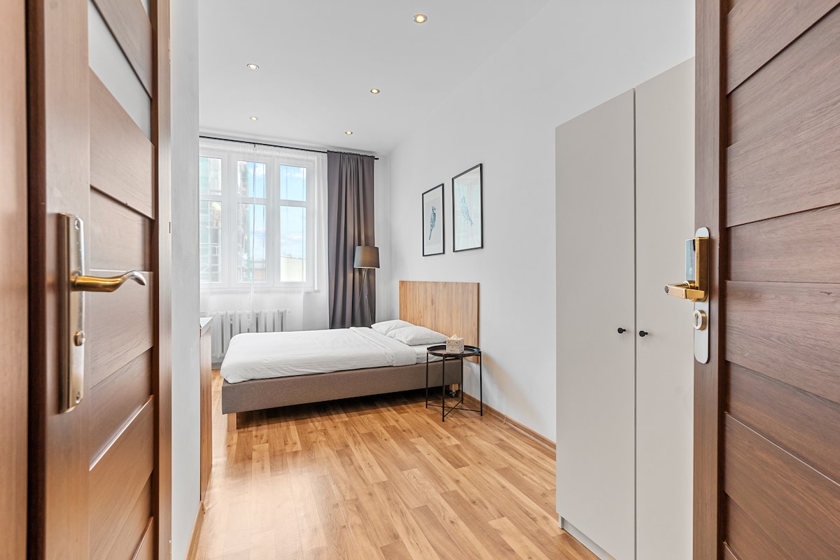 The studio features a spacious sleeping area with a bed positioned against a light wooden headboard. Natural light floods in through large windows, illuminating the warm wooden flooring. A small bedside table sits beside the bed, and a lamp is positioned nearby. A wardrobe stands against the wall.