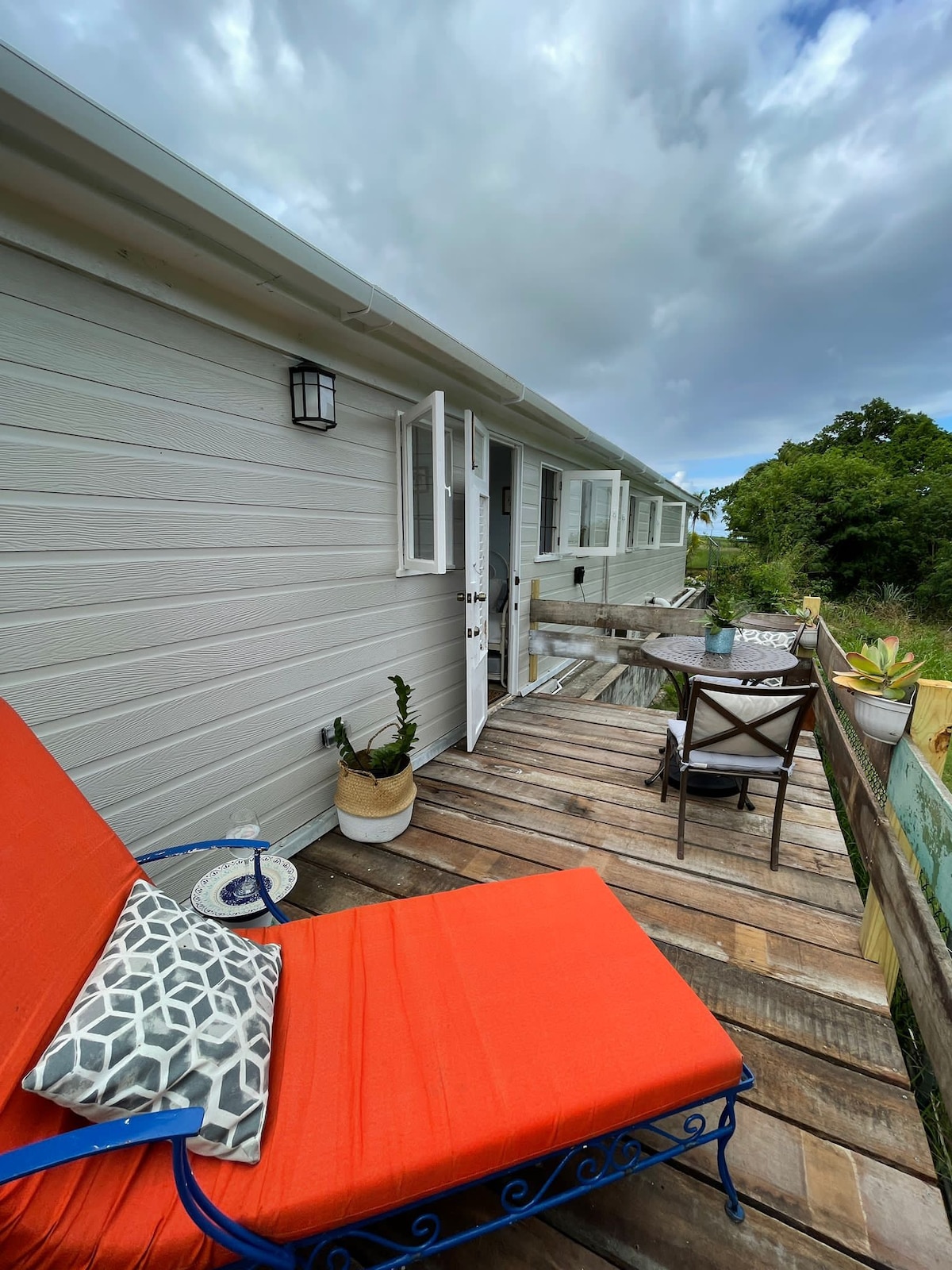 An outdoor seating area is featured with an orange lounge chair positioned on a wooden deck. A small table and a chair are visible nearby, surrounded by greenery and plants. Natural light enhances the space, with open double doors leading into the interior.