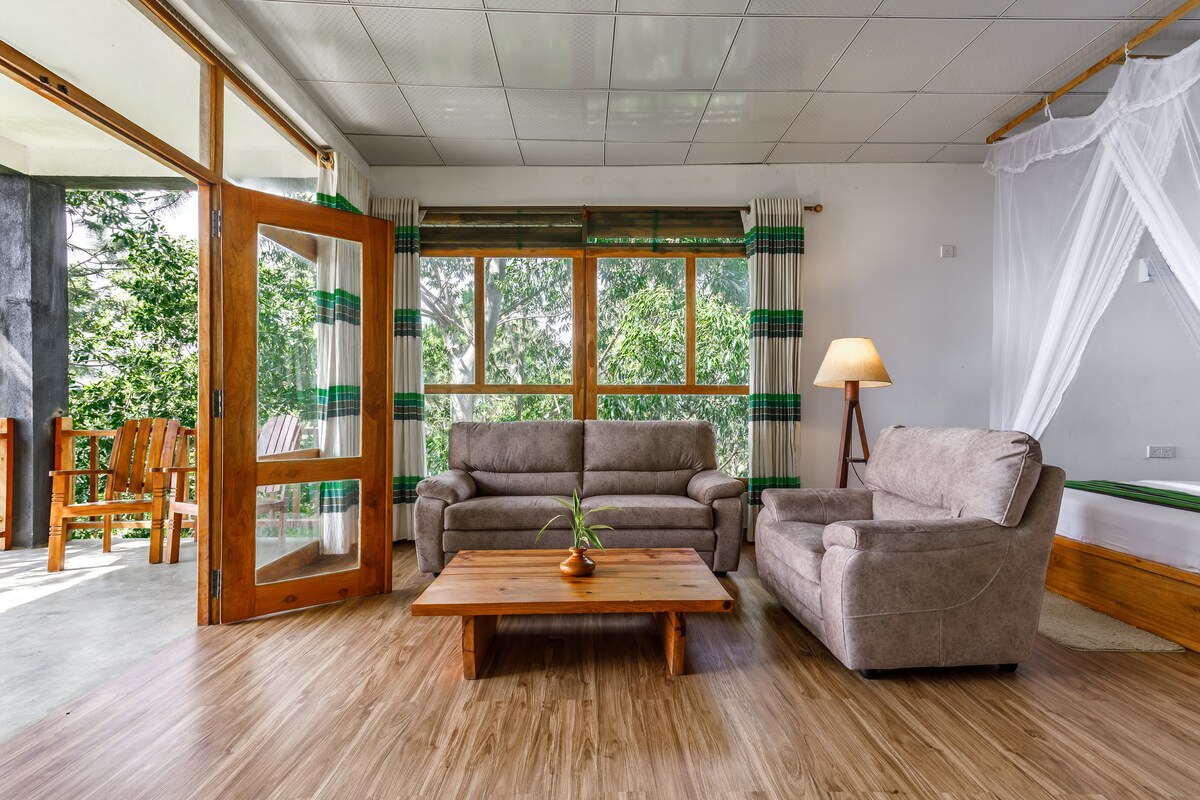 A living area is featured with a pair of light gray sofas positioned around a wooden coffee table. Large windows allow natural light to fill the space, revealing an outdoor view. A floor lamp stands beside the sofa, adding gentle illumination.
