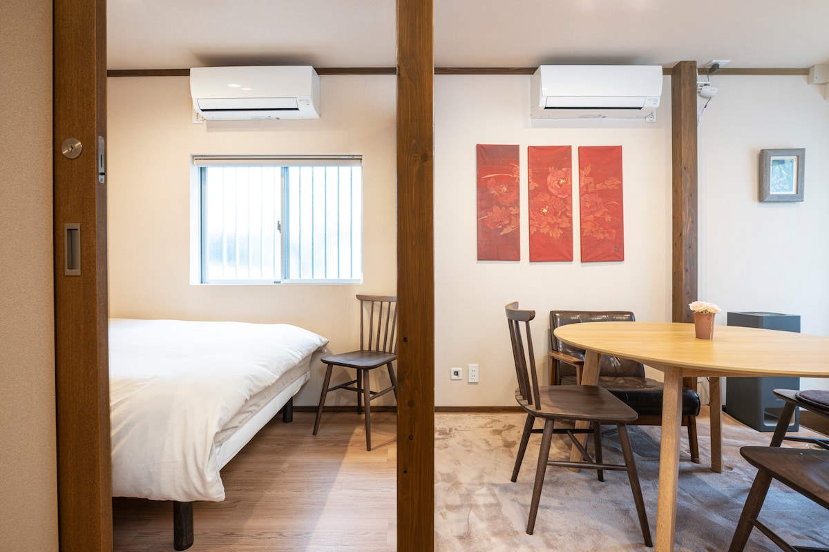 A welcoming bedroom area features a spacious bed, complemented by a round wooden dining table surrounded by chairs. Natural light enters through a window, with artwork adorning the walls, creating a cozy and functional living space.