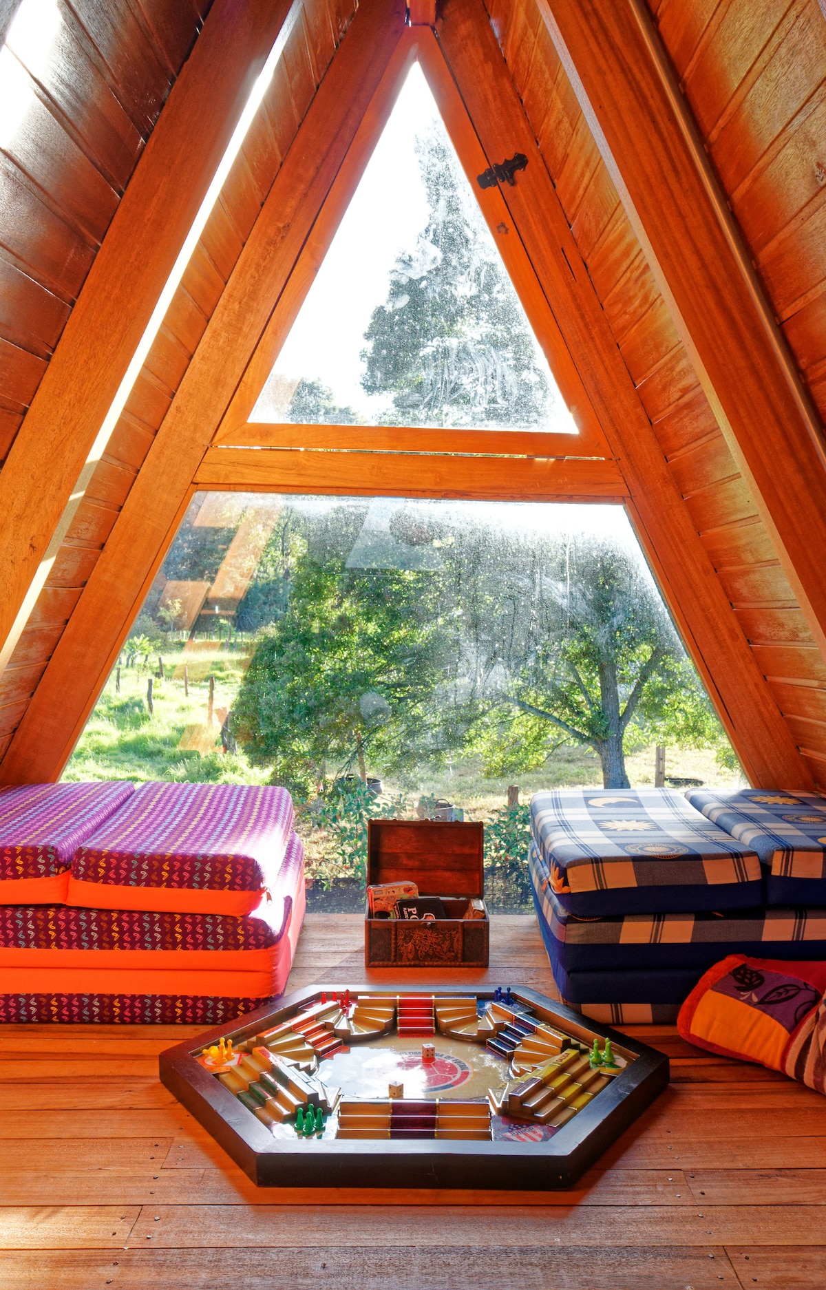 A triangular seating area features colorful mattresses arranged against wooden walls. A game board sits on a hexagonal table, with lush greenery visible through large windows that allow natural light to fill the space.