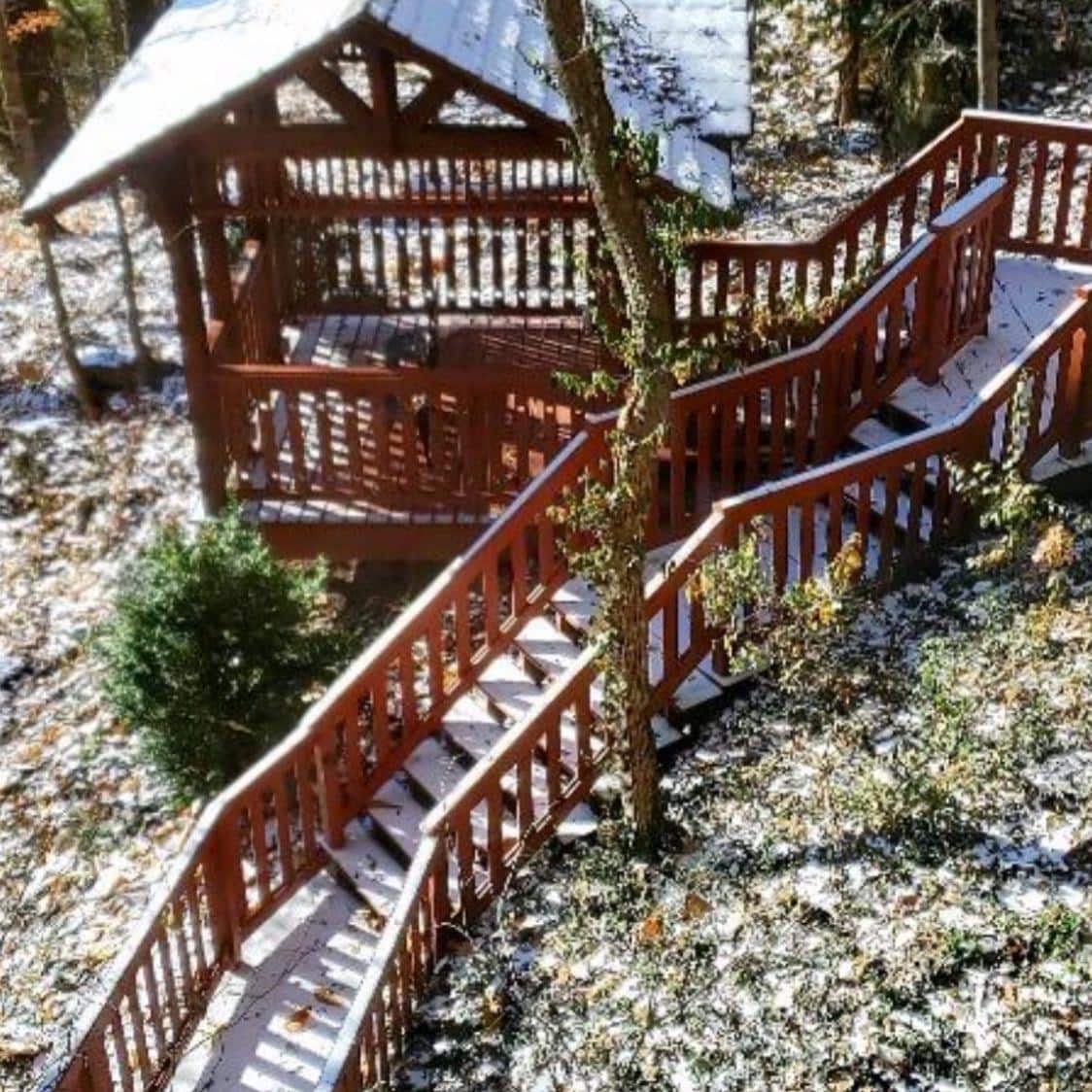 A multi-level wooden deck features winding staircases surrounded by a wooded landscape. Snow covers the steps and surrounding ground, creating a serene atmosphere. The natural elements enhance the privacy and seclusion of the outdoor space.
