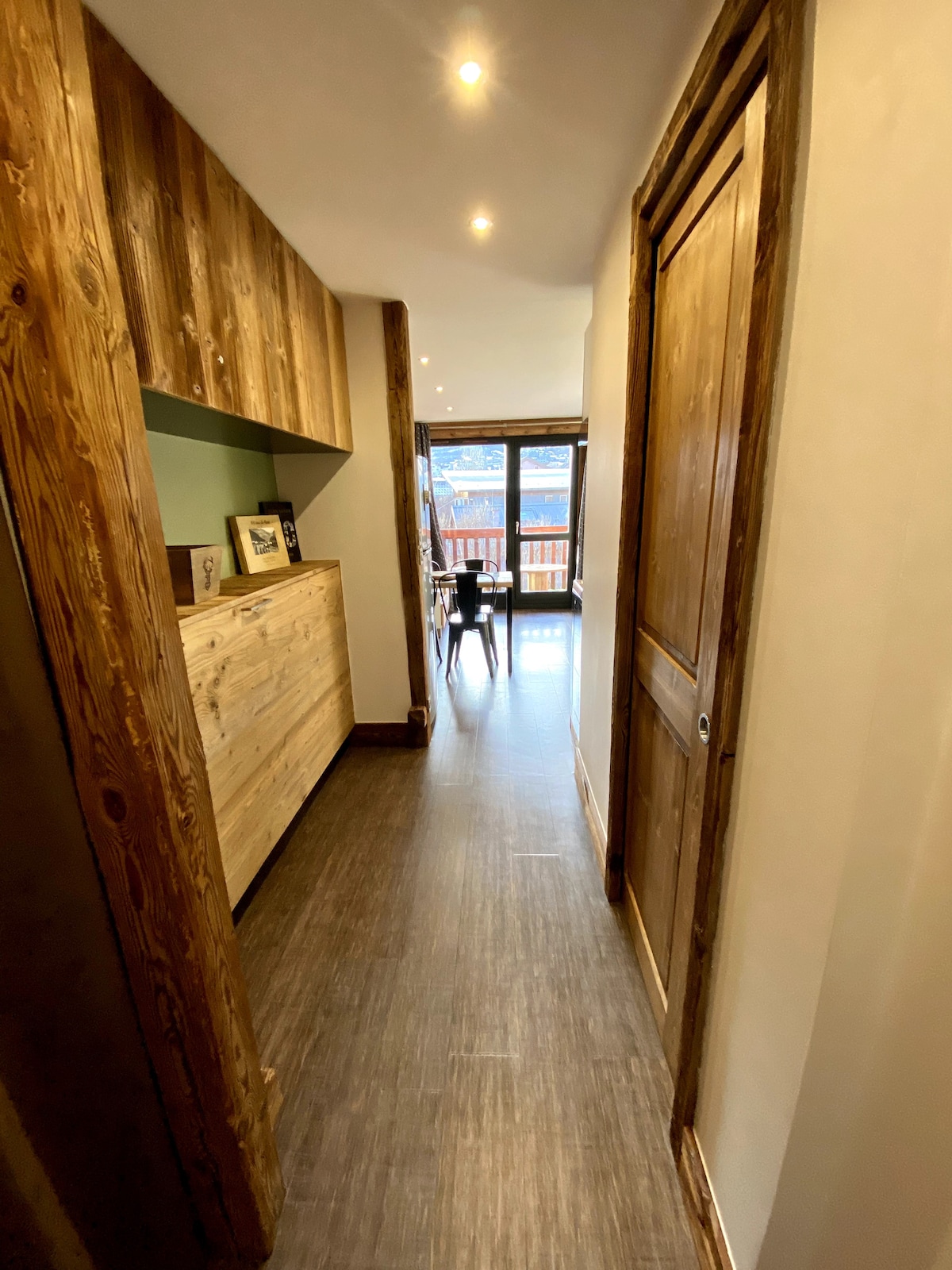 A narrow hallway is shown, lined with wooden walls and flooring. Natural light enters through a window at the end, casting a warm glow. A wooden sideboard and a door can be seen on one side, with a small dining table positioned at the far end.