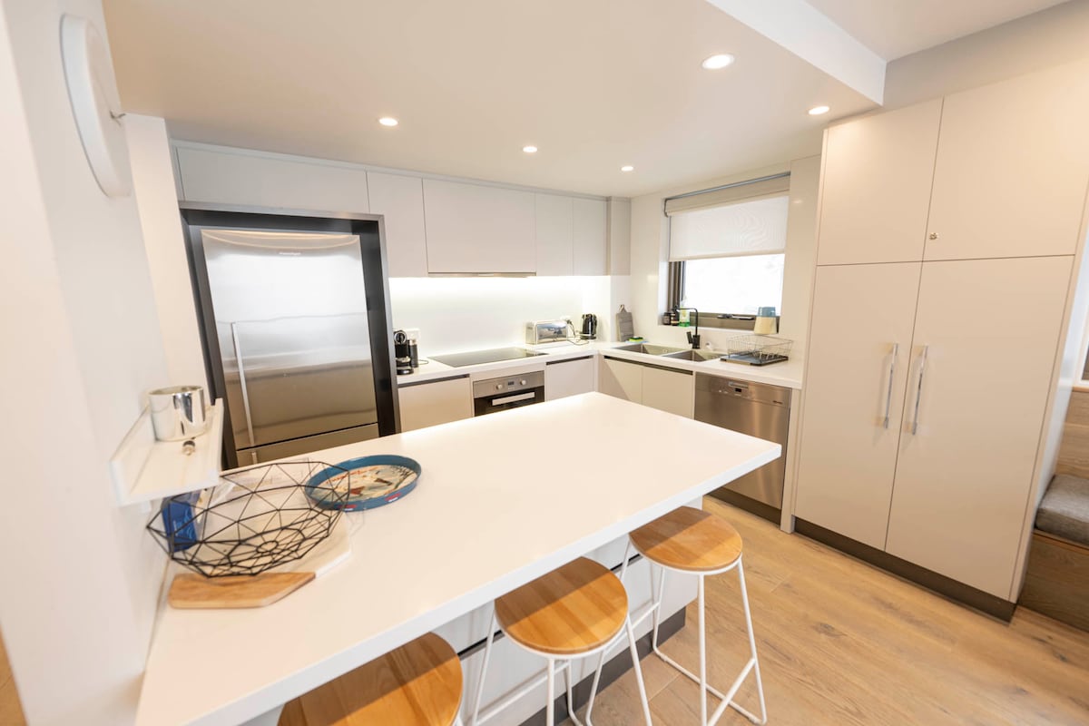A modern kitchen is equipped with sleek cabinetry and various appliances, including a refrigerator and dishwasher. A minimalist island with three wooden stools provides a casual dining space. Light is softly accentuated throughout the room, highlighting the stylish design and functional layout.