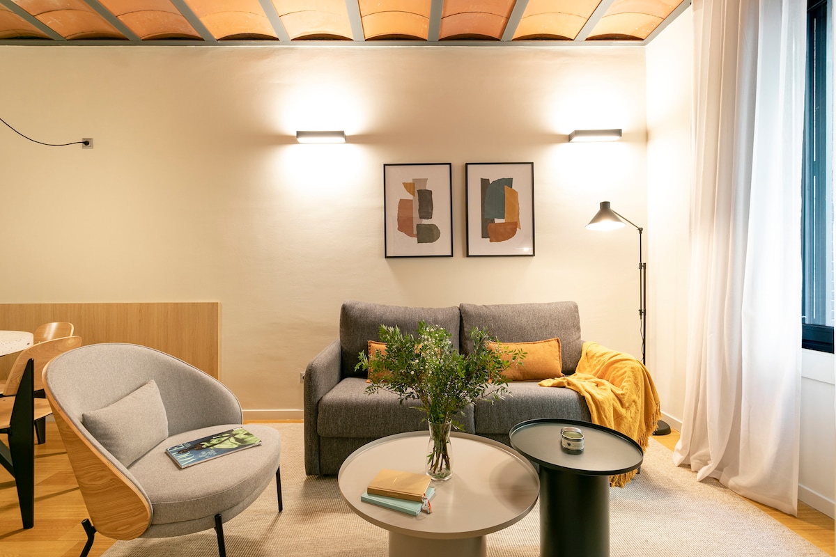 A cozy living area features a grey sofa adorned with a mustard throw, complemented by a round coffee table. Decorative wall art hangs above the sofa, while a floor lamp provides soft illumination. Natural light enters through a window draped with light curtains.