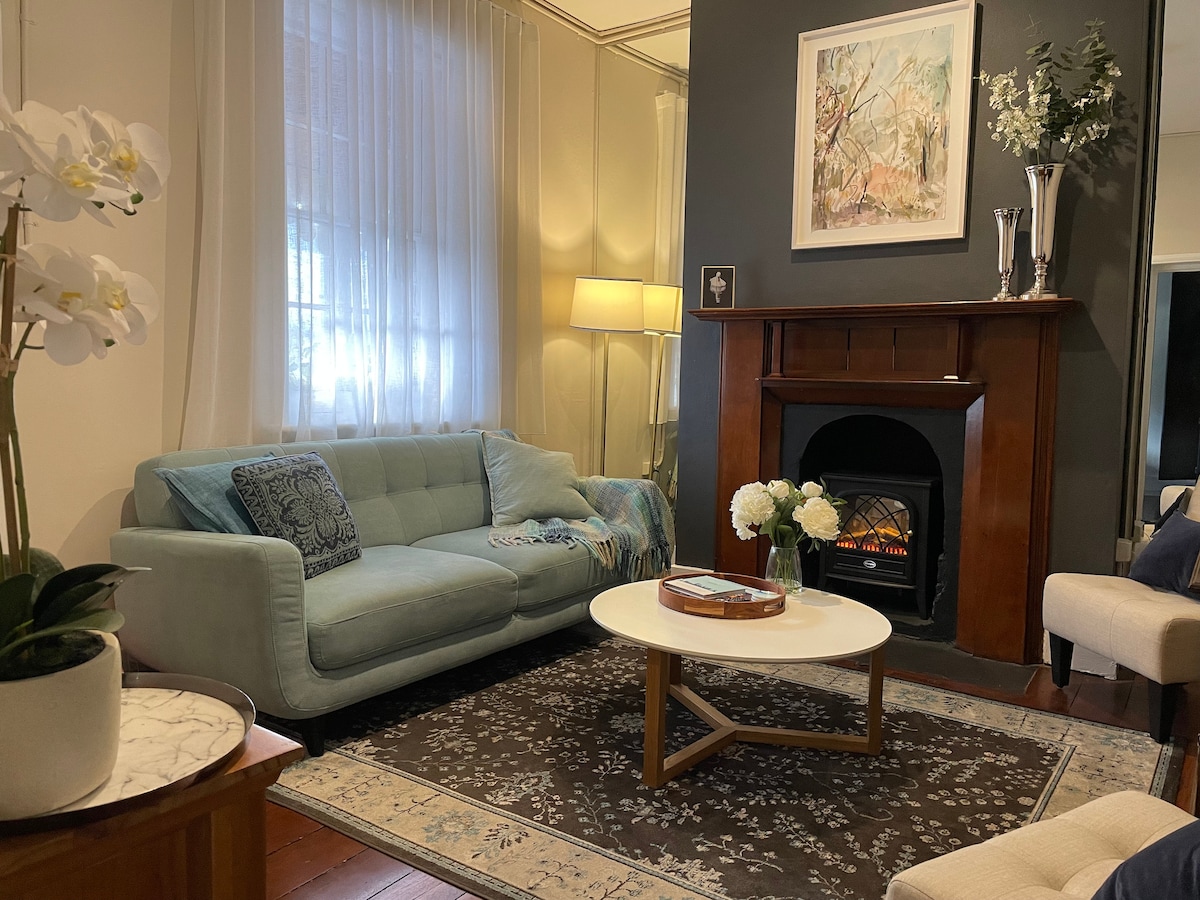 A cozy living room features a comfortable light blue sofa and a round coffee table arranged on a patterned rug. Natural light filters through sheer curtains, illuminating the space. A decorative fireplace adds charm, complemented by artwork and fresh flowers.