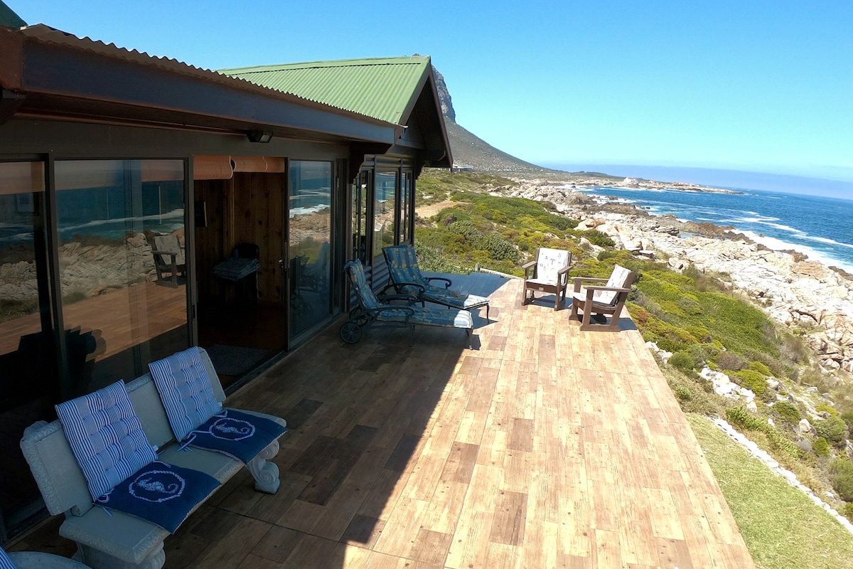 The image displays a sunlit wooden deck with chairs and loungers overlooking the ocean. Large windows reflect the scenic coastline, with lush greenery and rocky outcrops visible in the background. A coastal mountain can be seen in the distance under a clear blue sky.