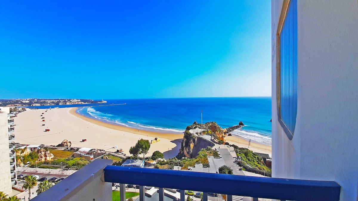 A view from the balcony showcases the expansive sandy beach and the turquoise ocean. The horizon meets a clear blue sky, with subtle coastal features visible, enhancing the natural beauty of the setting.
