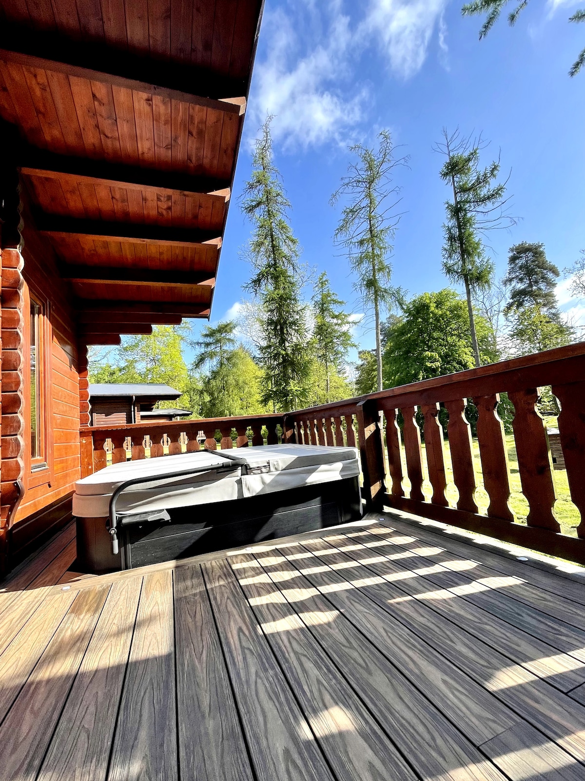 A spacious decking area is presented, featuring a luxury hot tub positioned in the sunlight. Wooden railings surround the deck, complemented by a backdrop of tall trees and clear blue skies, creating a serene and inviting outdoor space.