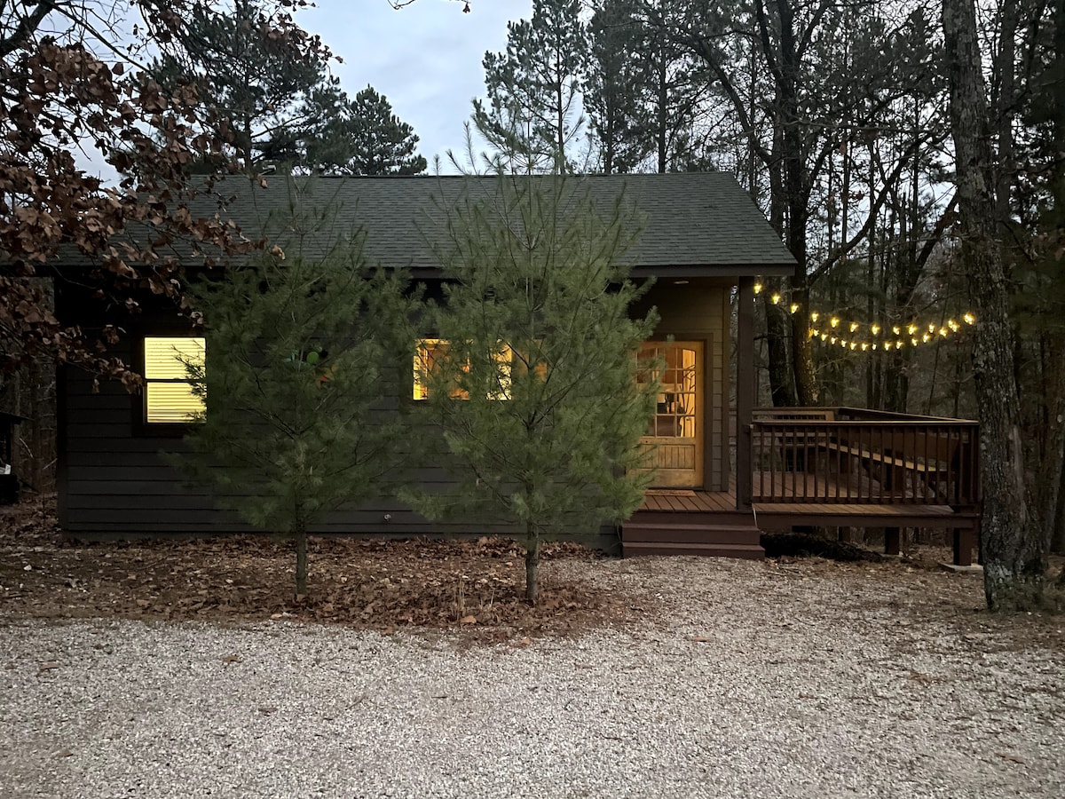 A cozy cabin sits among the trees, featuring a wooden deck with string lights adding soft illumination. The entrance is framed by lush greenery, and large windows reveal warm light from within, complementing the natural surroundings.
