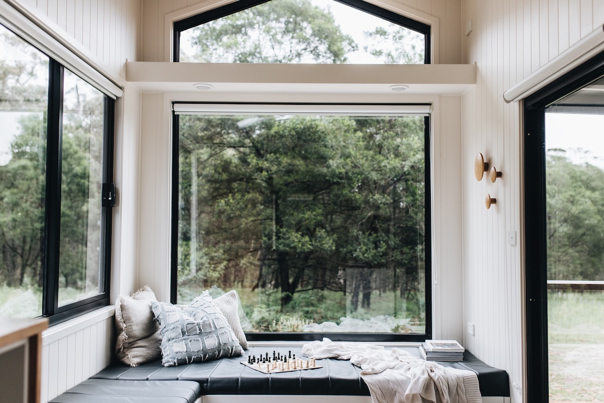 A cozy seating area features large windows that provide a view of the surrounding bushland. A comfortable black bench seat is adorned with soft pillows and a textured blanket. Natural light fills the space, highlighting the wooden accents and creating a serene atmosphere.