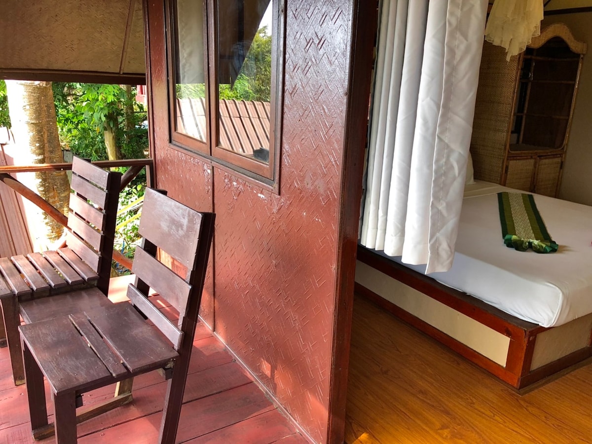 A wooden balcony presents two chairs, offering a view of the surrounding tropical greenery. A spacious bed is positioned inside the bungalow, partially visible through a window covered by light curtains. Warm wooden flooring enhances the inviting atmosphere of the retreat.