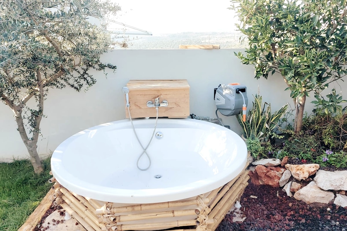 An outdoor bathtub is featured on a wooden base surrounded by natural stones. Lush greenery and a flowering bush provide a serene backdrop, while distant views of the landscape are observable in the background. A sleek faucet is mounted at the side of the tub.